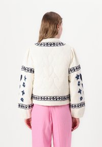 Quiltet cremet jakke med mønstret marineblå broderi på skuldre og ærmer. Har puffede ærmer og ribbede manchetter, samt et cropped design.