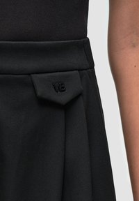 Černá skládaná sukně s výraznou nášivkou se znaky "WB" v černé barvě. Vyrobena z texturovaného materiálu s upraveným designem.