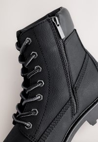 Čierna kožená čižma so štruktúrovaným povrchom, kovovými okami a sivými šnúrkami. Má bočný zips a pevný podrážkový vzor.