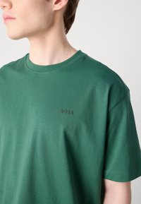 Zelené bavlněné tričko s designem kulatého výstřihu. Má krátké rukávy a jemné černé logo 'BOSS' na hrudi. Měkká textura.