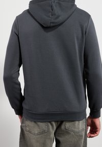Persona vestida con una sudadera gris oscuro lisa y jeans gris desvanecidos, vista desde atrás contra un fondo claro liso.