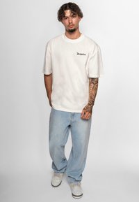 Tricou din bumbac crem, supradimensionat, cu text negru "Dropsize", asortat cu blugi de denim albastre deschise și teniși gri. Design simplu, croială lejeră.