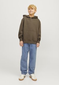 Sudadera con capucha marrón y bolsillo frontal, combinada con jeans de denim azul claro y zapatillas beige con rayas verdes y negras.