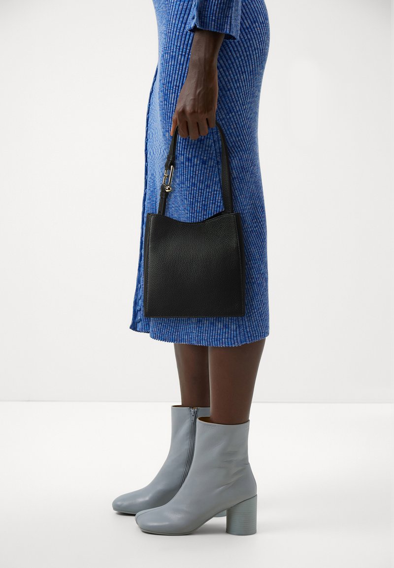 Black leather handbag with a curved top, held by a hand. The person wears a blue ribbed dress and light gray ankle boots with a block heel.