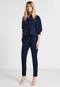 Femme debout les mains dans les poches, portant un blouse marine à petits motifs blancs, un pantalon assorti et des sandales à talons blancs.