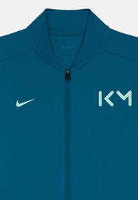 Μπλε αθλητικό μπουφάν με φερμουάρ, που διαθέτει το λευκό σήμα της Nike στα αριστερά και τα λευκά αρχικά "ΚΜ" στο δεξί στήθος.