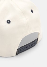 Casquette en toile blanche avec une sangle ajustable bleu foncé, des coutures contrastantes et une visière plate. Équipée de trous d'aération pour la respirabilité.
