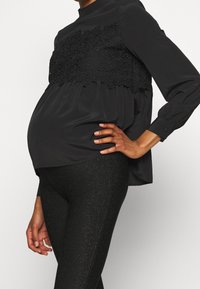 Czarna bluzka ciążowa z koronkowym wykończeniem w pasie, długimi rękawami i luźnym krojem, zestawiona z błyszczącymi czarnymi legginsami.