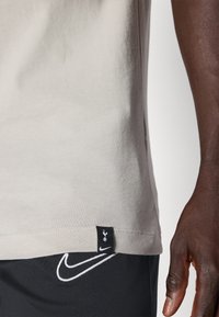 Camiseta de manga corta gris claro con una textura suave, presenta una etiqueta negra con un logotipo en el costado y un tejido oscuro debajo que muestra un emblema de Nike.