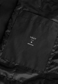 Étiquette de vêtement en tissu noir cousue, affichant la marque NEXT, taille M, et le texte "Made in China".