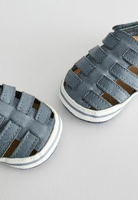 Grå vävda sandaler med stängd tådesign, vita ränder längs sålen och en slät yta. Har öppna utskärningar för andningsförmåga.