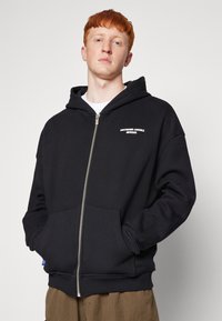 Black zip-up hoodie made from textured fabric, featuring a front kangaroo pocket, ribbed cuffs, and a white logo on the left chest.