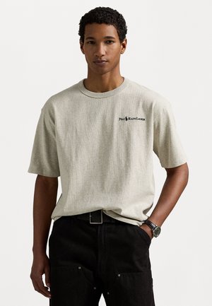 Tricou gri cu mânecă scurtă și guler rotund, având un mic logo negru pe partea stângă a pieptului; asortat cu pantaloni negri și o curea neagră.