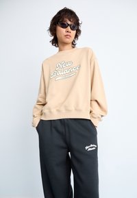 Sweat-shirt beige avec le logo blanc "New Balance Athletics", accompagné d'un pantalon de survêtement noir avec un logo brodé blanc. Tissu doux, coupe décontractée.