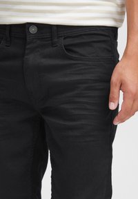 Črne jeans hlače iz jeansa z oprijeto zasnovo, z zapenjanjem na gumbe, sprednjimi žepi in rahlim bledenjem. Tekstura tkanine je videti gladka.