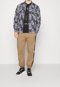 Marinblå skjorta med blommönster, svart t-shirt med grafik, beige joggers med sidosträck, och svarta sneakers med vita detaljer.