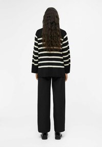 Schwarz-cream gestreifter, übergroßer Pullover mit langen Ärmeln und seitlichen Schlitzen, kombiniert mit schwarzen, weiten Hosen und glänzenden schwarzen Schuhen.