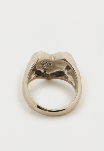 Zilverkleurige metalen ring in de vorm van lippen, bekeken van binnenuit, met kleine transparante steentjes die een letter vormen.