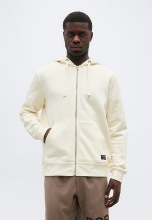 Muž v krémovej mikine na zips so sťahovacími šnúrkami a hnedých nohaviciach, stojaci na jednoliatej svetlej pozadí.