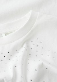T-shirt en coton blanc avec un col rond et des accents de strass en argent dispersés sur le devant. Texture douce visible.