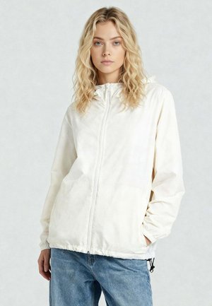 Jonge vrouw met golvend blond haar, gekleed in een witte hoodie met rits en blauwe jeans, staand tegen een effen lichte achtergrond.