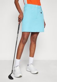 Žena v světle modré golfové sukni a bílých teniskách drží golfovou hůl a stojí před jednoduchým pozadím.