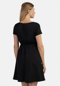 Rochie neagră cu mânecă scurtă, cu o croială lejeră, talie elastică și fustă fluida. Material neted și tiv până la genunchi. Vedere din spate.
