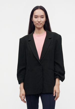 Jonge vrouw met lang donker haar, die een oversized zwarte blazer draagt, een roze top en marineblauwe broek tegen een effen witte achtergrond.