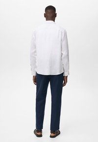 Valkoinen pellavainen pitkähihainen paita, jossa on avara kaulus, yhdistettynä tummansinisiin slim-fit-housuihin, mustat sandaalit jalassa; siisti muotoilu ja kevyt rakenne.