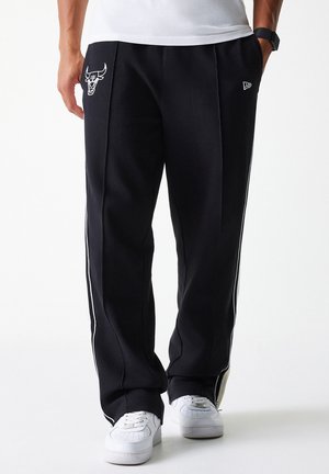 Black athletic pants with white side stripes, feature an embroidered logo on the left thigh, made from soft, textured fabric, styled for comfort.