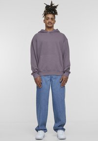 Felpa viola over-size con tasca frontale, abbinata a jeans in denim larghi e sneakers bianche. Lo sfondo è neutro e uniforme.