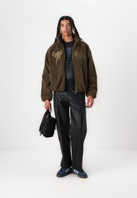 Olivgrön fleecejacka med dragkedja, prydd med tribalbroderi, i kombination med svarta fake läderbyxor och blå sneakers med ränder. Håller en svart väska.