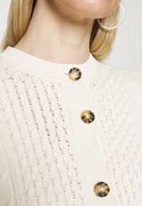 Cremfarbener Strickcardigan mit strukturiertem Muster, ausgestattet mit großen runden Schildpattknöpfen vorne und einem Rundhalsausschnitt.