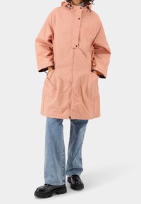 Osoba oblečená v lososově růžové mikině s kapucí, s rukama v kapsách, světlých modrých džínách a černých masivních botách, stojící proti jednobarevnému pozadí.