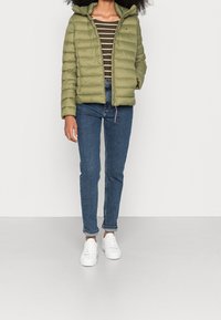 Giacca imbottita verde oliva con cappuccio, dotata di zip frontale e cuciture orizzontali, indossata sopra una maglietta a righe a maniche lunghe e jeans blu.