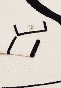 Krém szövet textúrált bordázattal, fekete kiegészítőkkel és dekoratív kristálygombbal. Szerkezettartó gallérral és mandzsettákkal rendelkezik.