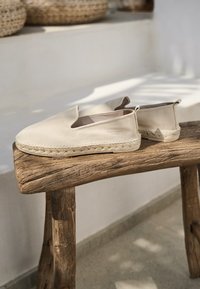 Espadrillas beige in tessuto morbido con suola in juta intrecciata, caratterizzate da una superficie liscia e un design minimalista, esposte su un supporto in legno.