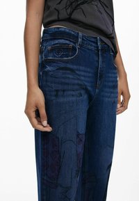 Jeans en denim bleu foncé avec une coupe décontractée, des détails graphiques subtils sur les jambes et un design classique à cinq poches avec des coutures orange.