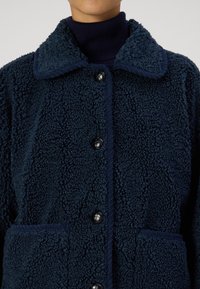 Granatowa polarowa kurtka z teksturowaną powierzchnią, dużym kołnierzem, czarnymi guzikami oraz dwiema przednimi kieszeniami z kontrastowym wykończeniem.