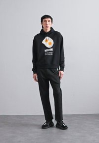 Schwarzer Hoodie mit einem Grafikdesign eines Spiegeleis und "MOSCHINO"-Schriftzug; kombiniert mit schwarzen Hosen und schwarzen Schuhen; glattes Material, normale Passform.