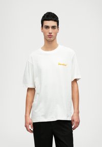 Camiseta blanca de algodón con mangas cortas y escote redondo. Presenta el logo "Paradisø" en amarillo en la parte izquierda del pecho.