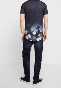 Svart kortärmad t-shirt med blommig gradientdesign vid fållen, tillsammans med mörka jeans och svarta sneakers, sedd bakifrån.