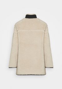 Krämfärgad, texturerad shearlingkappa med hög krage, svarta detaljer och rak klippning. Sömmen över ryggen ger struktur.