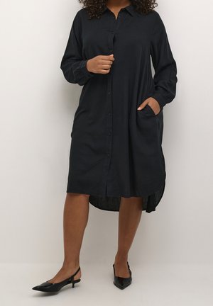 Femme portant une robe chemise noire à manches longues avec un ourlet asymétrique, une main dans la poche, associée à des chaussures slingback noires à talons bas.