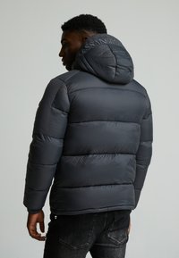 Mørkegrå pufferjakke med hette, quiltet design, og brodert merkevarelogo på baksiden. Teksturerte stoff med en glatt finish.