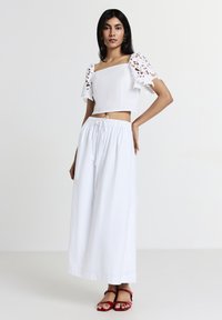Femme portant un crop top blanc à manches en dentelle, une longue jupe blanche avec taille à cordon et des sandales rouges à bout ouvert, se tenant contre un fond uni.