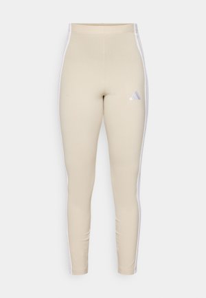 Béžové legíny s vysokým pásom, biele bočné pruhy a vyšívané logo so tromi diagonálnymi pruhmi v purpurovej a modrej farbe.