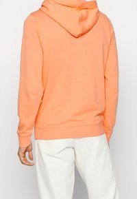 Felpa arancione con cappuccio regolabile, orlo e polsini a coste, realizzata in un tessuto morbido. Indossata con pantaloni chiari, vista posteriore.
