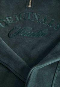 Ciemnozielona bluza z designem na ćwierć zamek. Posiada wytłoczone napisy "ORIGINALS" i "Studio" w dopasowanym odcieniu. Miękka tekstura.
