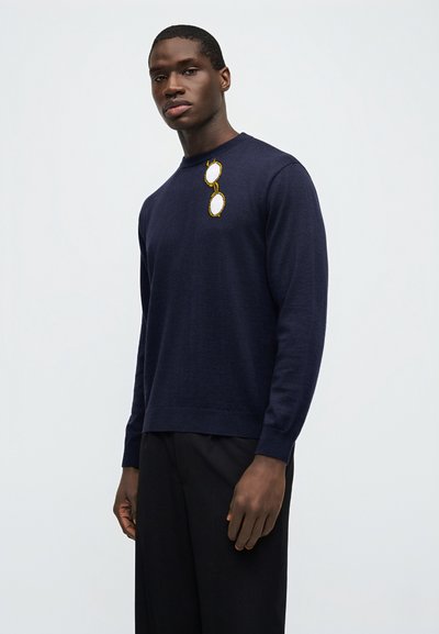 Άνδρας που φοράει μπλε σκουρόχρωμο πουλόβερ με κεντημένα γυαλιά ηλίου στο στήθος και μαύρο παντελόνι, στέκεται μπροστά σε απλό φωτεινό φόντο.